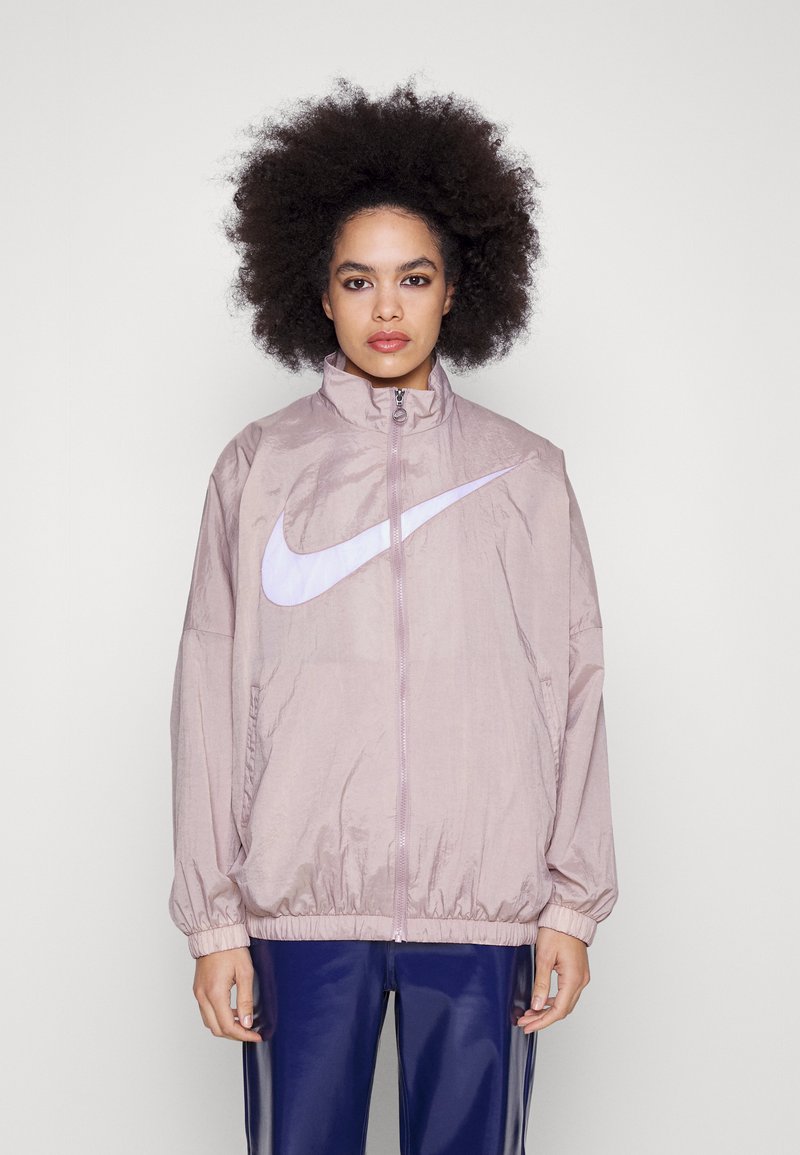 Ljusrosa Nike vindjacka med hög krage, långa ärmar, elastiska muddarna och en stor vit swoosh-logotyp på framsidan.