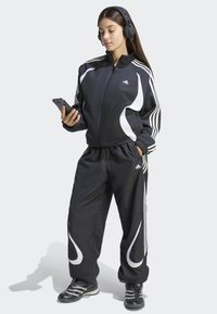 Crni trenirka s bijelim detaljima, uključujući jaknu na patentni zatvarač i uske hlače. Osoba nosi crne slušalice i drži pametni telefon.