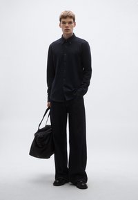 Fekete, hosszú ujjú gombos ing, széles sötét nadrággal párosítva. A modell egy fekete táskát tart a kezében. Simaságú anyag és egyszerű kivitelezés.