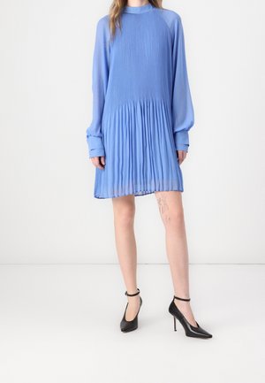 Kobieta w jasno-niebieskiej plisowanej sukience z długim rękawem, w połączeniu z czarnymi szpilkami na kostkę, stojąca na tle jednolitego białego tła.