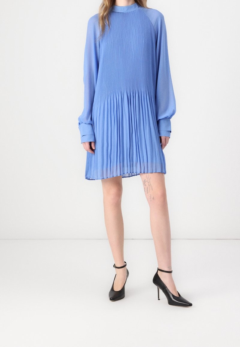 Femme portant une robe plissée à manches longues bleu clair, associée à des escarpins noirs à bride cheville, debout devant un fond blanc uni.