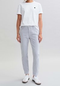 Bílý tričko s malým černým srdcem na hrudi, spárované s světle šedými slim-fit chino kalhotami a bílými teniskami s hnědými detaily.