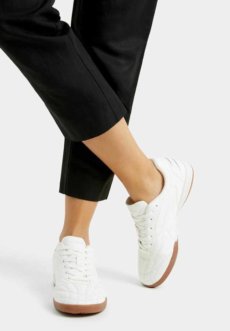 Białe sneakersy z chropowatą skórzaną cholewką, gumową podeszwą i sznurowanym wzorem. Buty mają detale w postaci przeszyć oraz niski profil.