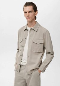 Beige lightweight jacket with a collar, two chest pockets, button closure, and long sleeves. Worn over a white t-shirt and matching pants.