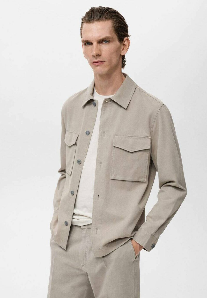Beige lightweight jacket with a collar, two chest pockets, button closure, and long sleeves. Worn over a white t-shirt and matching pants.