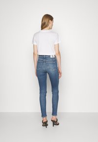 Modré slim-fit džíny s středním pasem, zdobené designem zadní kapsy a štítkem s logem CK. Oblečené s bílým zkráceným tričkem.