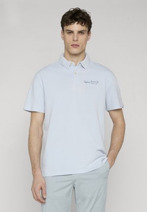 Ung mann med krøllete hår som har på seg en lys blå polotrøye med krage og lyse bukser, står med den ene hånden i lommen mot en ensfarget bakgrunn.