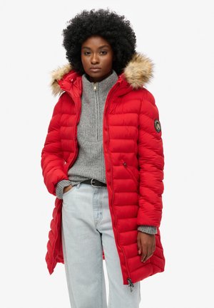 Red puffer coat with a fur-lined hood, featuring a zip front, quilted design, and logo patch on the sleeve, worn over a gray sweater.