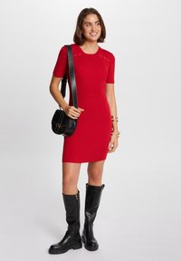 Rode geribbelde jurk met korte mouwen, ronde halslijn en gedetailleerde schouderaccenten. Gecombineerd met kniehoge zwarte leren laarzen en een zwarte handtas.