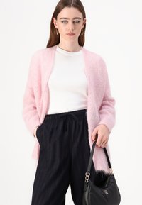 Lys rosa, fluffy cardigan med åpen front, båret over en hvit topp; kombinert med svarte stripete, løstsittende bukser og en svart håndveske.