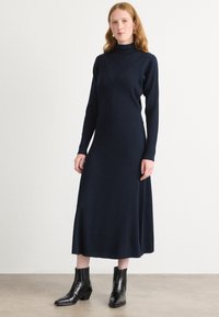 Vestido de punto acanalado en color azul marino con cuello alto, mangas largas y dobladillo acampanado; combinado con botas negras hasta el tobillo. Textura suave y corte ajustado.