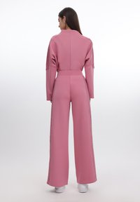 Rosa jumpsuit med vide ben, teksturerte blonder langs sidene og en tettsittende midje. Glatt stoff og lange ermer fullfører designet.