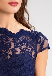 Nainen, jolla on päällään tummansininen pitsimekko, jossa on kukka- ja kiehkura-akuvioita, läpinäkyvä yläosa ja lyhyet hihat, sekä luonnolliset, laineikkaat ruskeat hiukset näkyvissä.