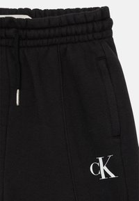 Svarta träningsoveraller i mjukt tyg, med elastisk midja och snörning samt en vit logotyp nära fickan.