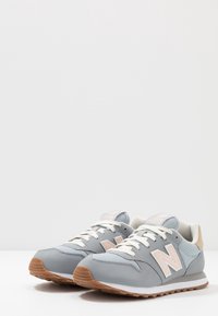 Par de zapatillas New Balance en gris y beige con cordones blancos y suelas de goma, colocadas lado a lado sobre una superficie blanca.