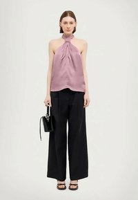 Kvinna i en ärmlös mauve halterneck-top, vida svarta byxor, svarta sandaler och håller en liten svart handväska.