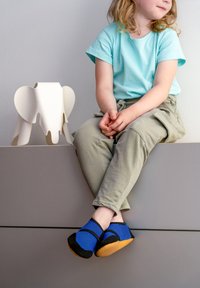 Het kinderschoeisel heeft een blauwe mesh bovenkant met zwarte accenten en een rubberen zool. Het staat naast een wit olifantvormig krukje.