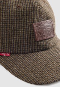 Brun tweedkeps med ett rutig mönster, med en brun läderpatch och en röd tagg på sidan. Slät textur och böjd skärm.