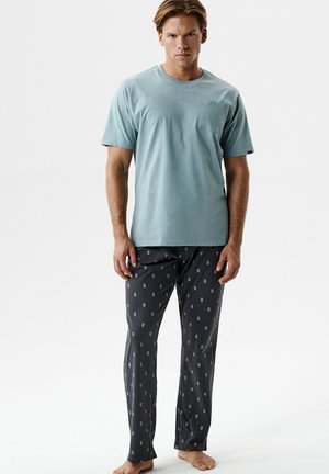 Mężczyzna w jasnobłękitnej koszuli z krótkim rękawem z kieszenią na piersi oraz czarnych, wzorzystych spodniach do piżamy, stojący boso na białym tle.