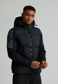 Μαύρο μπουφάν πούφ με ψηλό γιακά και κουκούλα. Διαθέτει κλείσιμο με φερμουάρ, πλαϊνές τσέπες και αντίθετα γκρίζα μανίκια. Λογοτύπο στο στήθος.