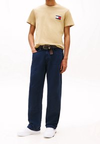 Camiseta de algodón beige con un parche de logotipo, combinada con pantalones de pierna ancha en navy y un cinturón marrón. Zapatillas blancas completan el conjunto.