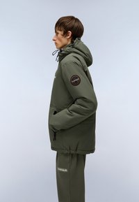 Oliven grøn hættetrøje med snørrebånd, stor forlommer og et rundt logo mærke på venstre ærme. Bæres med matchende sweatpants.