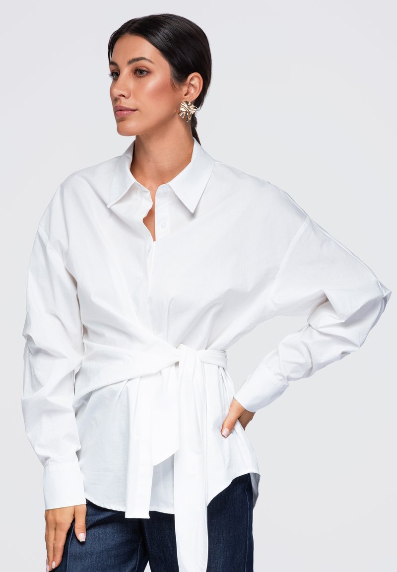 Femme aux cheveux foncés attachés en arrière portant une chemise blanche à manches longues boutonnée avec une ceinture à la taille et un pantalon foncé, posant avec la main sur la hanche, regardant à gauche.