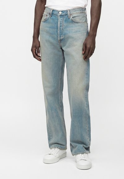 Homme portant un jean droit bleu clair et des baskets blanches, debout les mains détendues le long du corps sur un fond uni.
