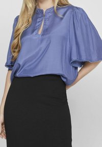 Lavendelfärgad blus med kort ärm, nyckelhalsringning och scallop-edgar, kombinerad med en figurnära svart kjol. Slät textur, avslappnad design.