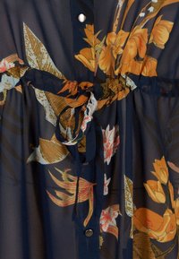 Blusa a fantasia floreale in tessuto trasparente blu navy con chiusura a bottoni e cintura annodata in vita. Presenta dettagli floreali gialli e arancioni.