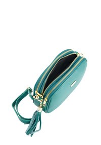 Tyrkisblå lædertaske med rem til at bære over skulderen, to lynlåslommer, gulddetaljer og en kvast som pynt. Glat tekstur og afrundet form.