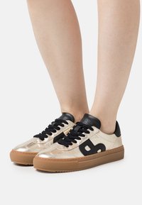 Gold metallic sneakers with black accents, featuring a textured finish, black laces, and a brown rubber sole. Logo detail on the side.