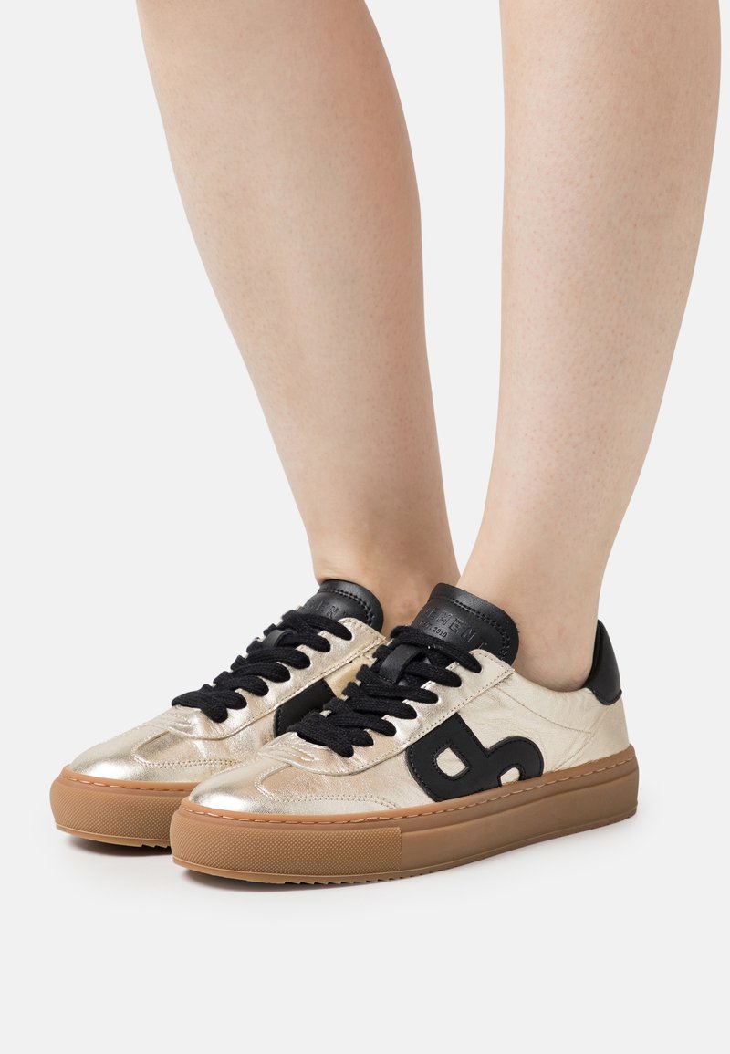 Gold metallic sneakers with black accents, featuring a textured finish, black laces, and a brown rubber sole. Logo detail on the side.