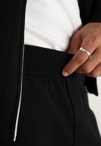 Hand with silver ring adjusting the ribbed waistband of black knit pants, wearing a white ribbed shirt and black zip-up sweater.
