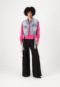 Gilet di denim sopra una felpa rosa brillante, abbinato a pantaloni neri larghi. Una piccola borsa rosa pende da una mano. Design semplice, vestibilità casual.