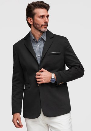 Homme barbu portant un blazer noir, une chemise bleue à motifs, un pantalon blanc et une montre-bracelet argentée, boutonnant son blazer, regardant sur le côté.