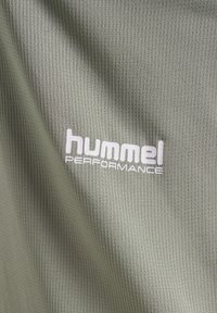 Olivgrönt, texturerat tyg med ribbad struktur. Har en tydligt synlig vit "hummel PERFORMANCE"-logotyp.