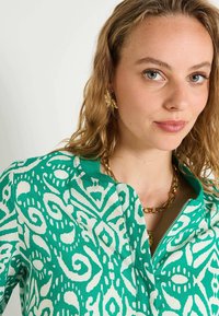 Camisa de patrones verde y blanca con una textura suave, que presenta un cuello con cremallera y un diseño de espiral audaz. Los accesorios dorados complementan el conjunto.