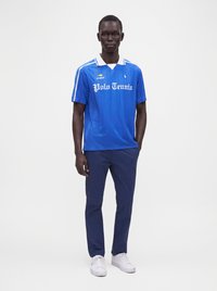 Homme debout portant un polo de tennis bleu, un pantalon bleu foncé et des baskets blanches sur un fond clair uni.