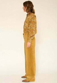 Blouse à manches longues couleur moutarde avec motif paisley, associée à un pantalon en velours côtelé taille haute à jambes larges de la même couleur. Chaussures plates.