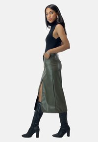 Top negro sin mangas combinado con una falda de cuero verde oliva de tiro alto con una abertura lateral, complementada con botas negras de tacón alto hasta la rodilla.