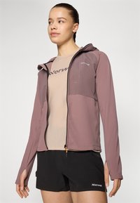 Veste zippée lilas avec un design ajusté, deux poches avant et une capuche, portée sur un t-shirt en maille beige et un short noir.