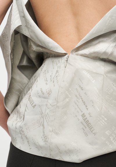 Zadnji del prikazuje delno zadrgan bež zgornji del z natisnjenimi vzorci besedila, ki razkriva zgornji del hrbta, v kombinaciji s temnimi hlačami.