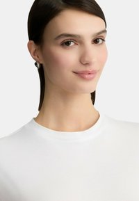 T-shirt bianco a maniche corte con scollatura rotonda. Il modello ha i capelli scuri e indossa un orecchino d'argento con un accento nero. Texture liscia.