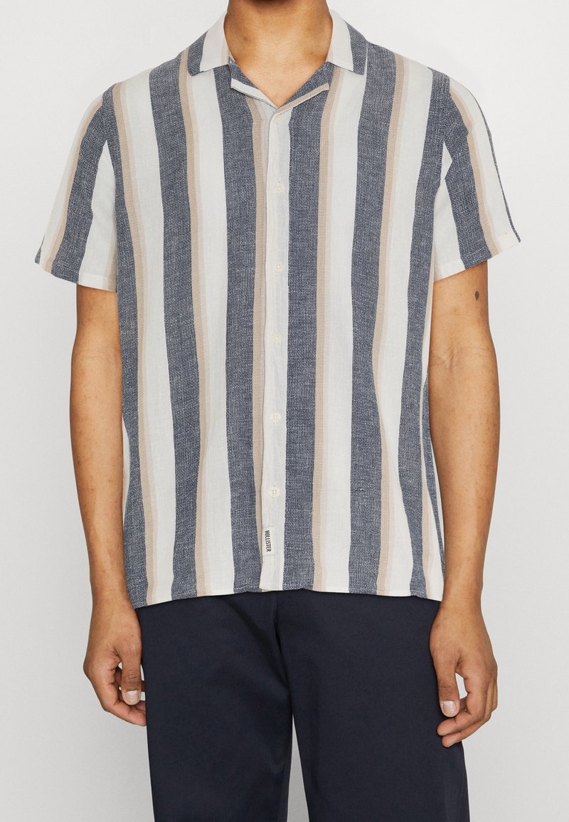 Koszula na krótki rękaw zapinana na guziki w pionowe paski w kolorach granatowym, beżowym i białym, wykonana z lekkiego materiału. Posiada projekt kołnierza typu camp.