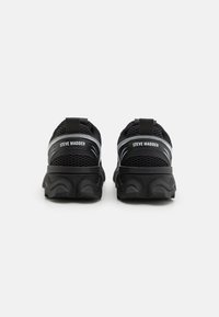 Zapatillas deportivas de malla negra con suelas de goma gruesa y un logotipo en el talón. Presentan una superficie texturizada y un diseño de suela curvada distintivo.
