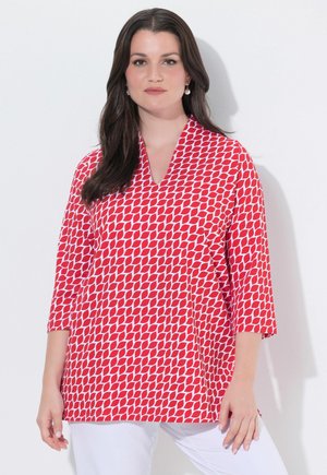 Kvinde med langt mørkt hår iført rød og hvid bluse med geometrisk mønster og trekvartlange ærmer samt hvide bukser, stående mod en hvid baggrund.