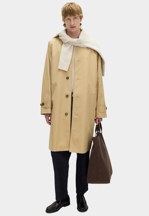 Mężczyzna w beżowym płaszczu typu trench, z lekkim swetrem zarzuconym na ramiona, ciemne spodnie, czarne buty, trzymający dużą brązową torbę na ramię na jednolitym tle.