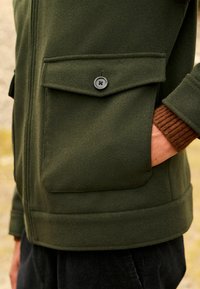 Hand in pocket of olive green jacket with large buttoned flap pocket, worn over brown ribbed sleeve and black pants.