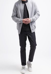 Grau, leichte Bomberjacke über einem strukturierten schwarzen Pullover, kombiniert mit dunklen Hosen und hellgrauen Sneakers vor einem schlichten Hintergrund.